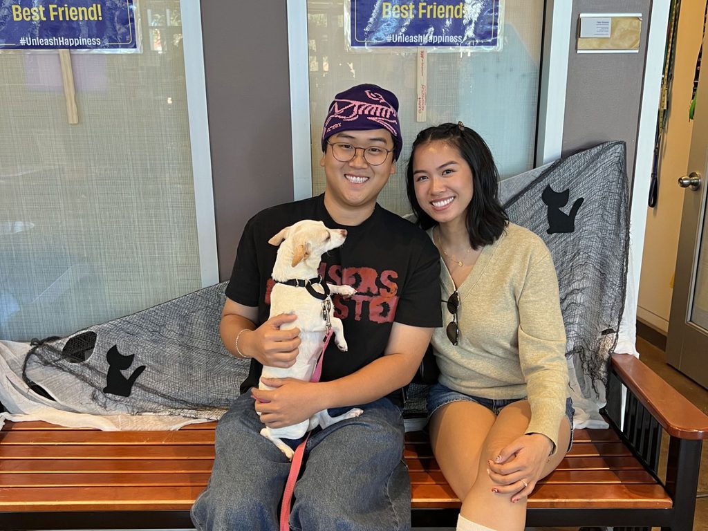 A smiling couple sits on a wooden bench holding a small white dog, with pet adoption signs and a glass wall in the background.