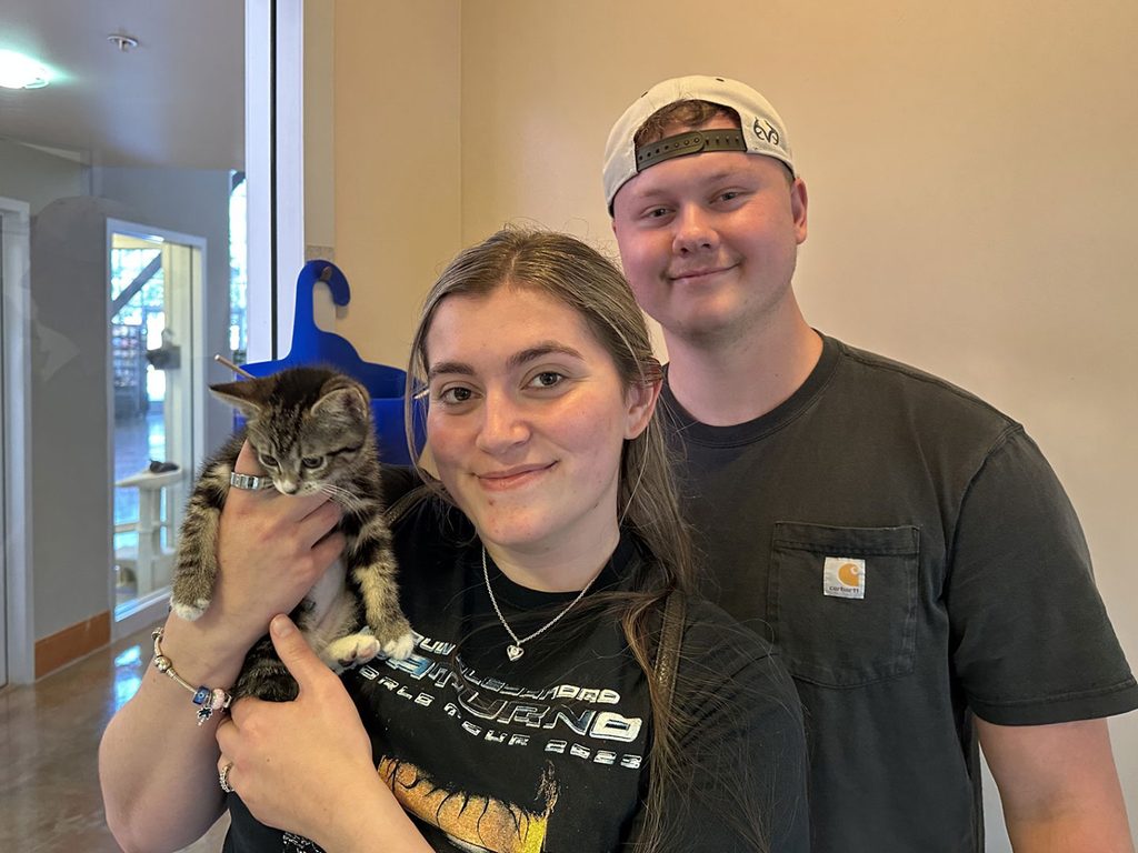 A woman holding a tabby kitten stands beside a man in a black shirt and white cap, with a doorway and hallway in the background.