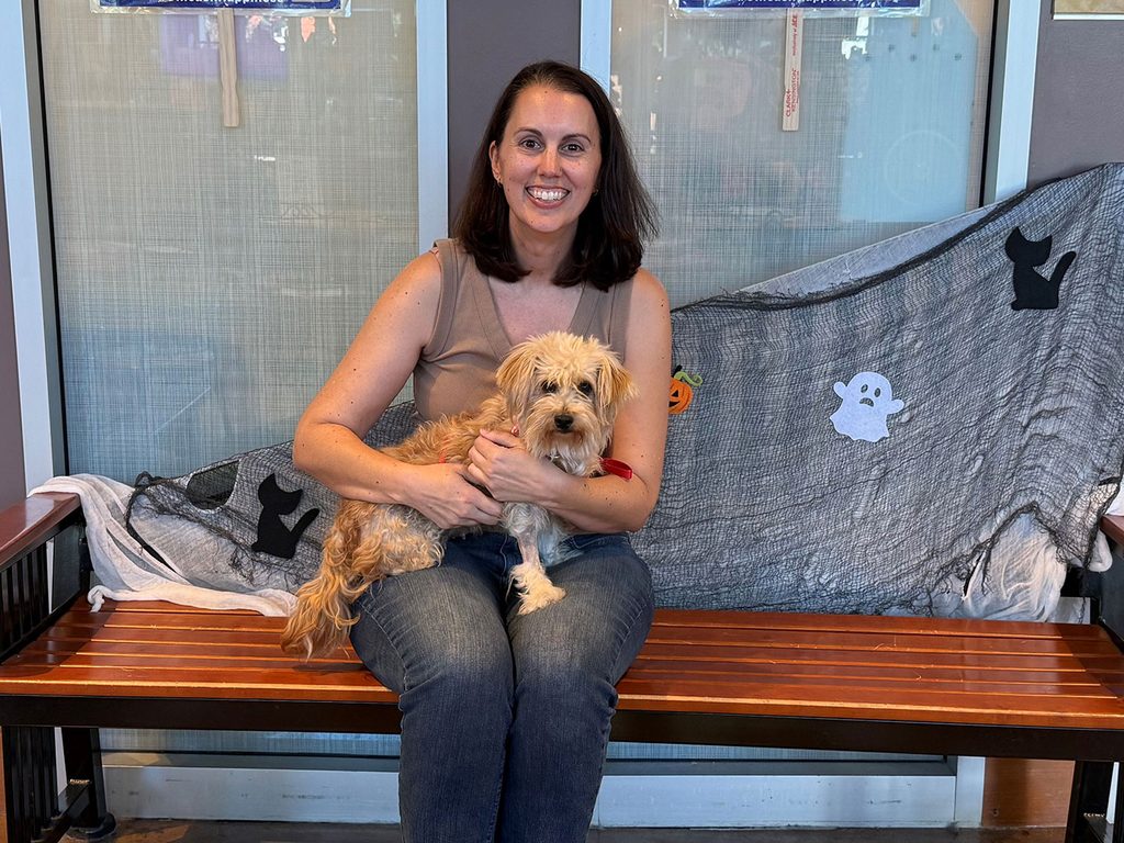 A woman sits on a wooden bench holding a small tan dog, with a gray blanket draped over the bench and glass doors behind them.