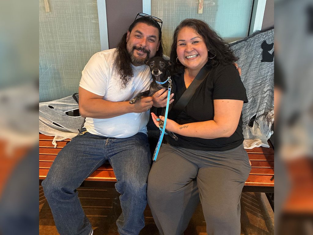 A man and woman sit on a bench holding a small black dog on a blue leash, with a blanket draped over the bench in the background.