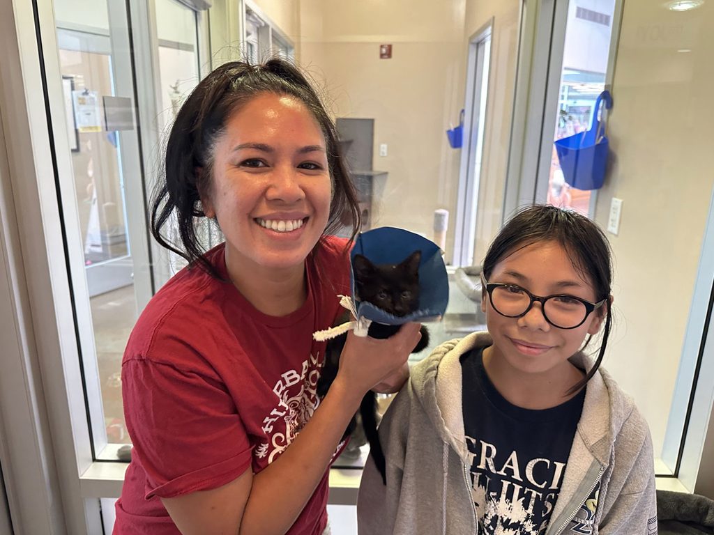 A woman in a red shirt holds a black cat wearing a blue cone, standing beside a girl in glasses and a gray hoodie in a bright indoor hallway.