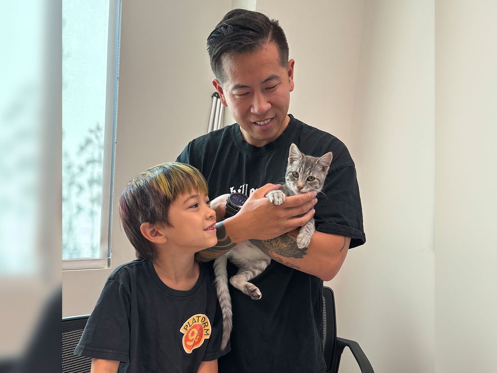 An adult man holds a gray tabby kitten while a young boy stands beside him, with a window and light-colored wall in the background.