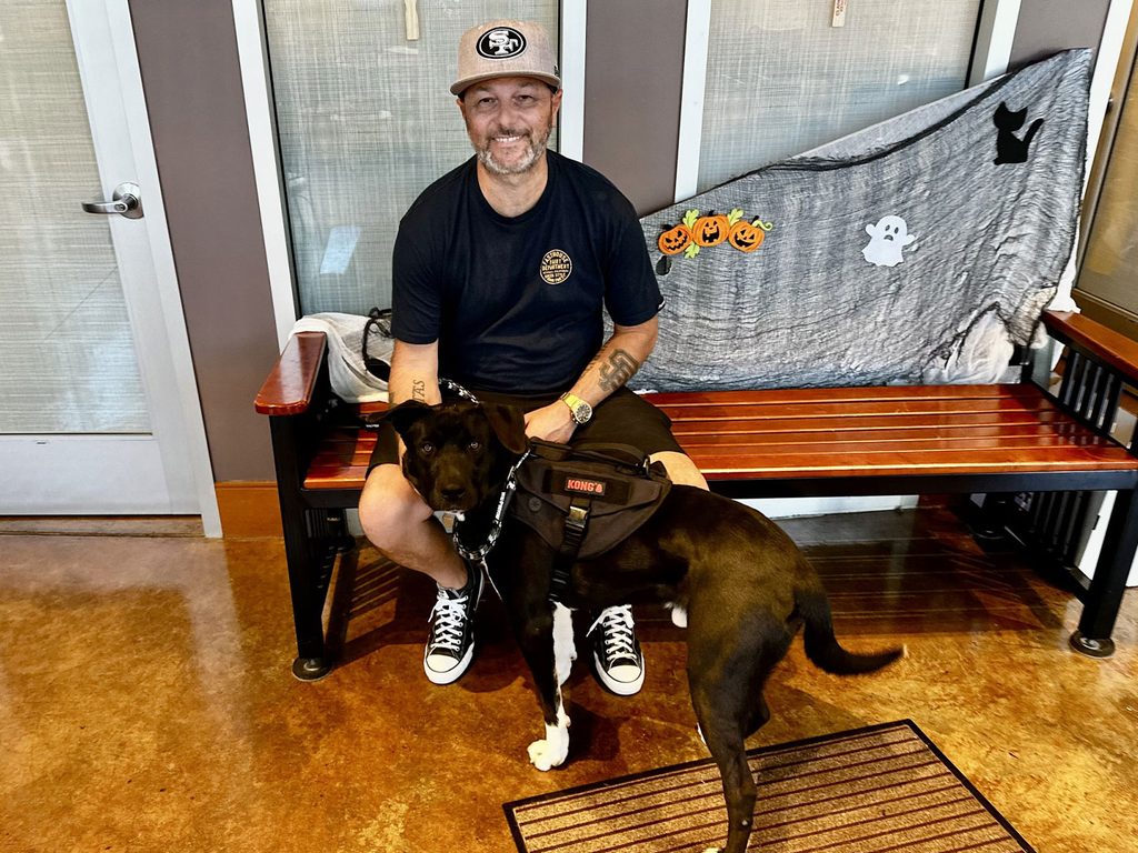 A man wearing a cap sits on a bench with a black dog in front of a gray Halloween-decorated backdrop.