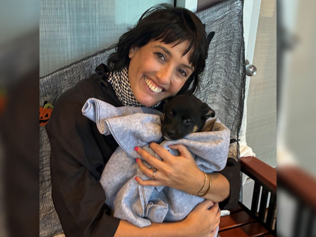 A woman with dark hair holds a small black puppy wrapped in a gray blanket while sitting in a chair indoors.