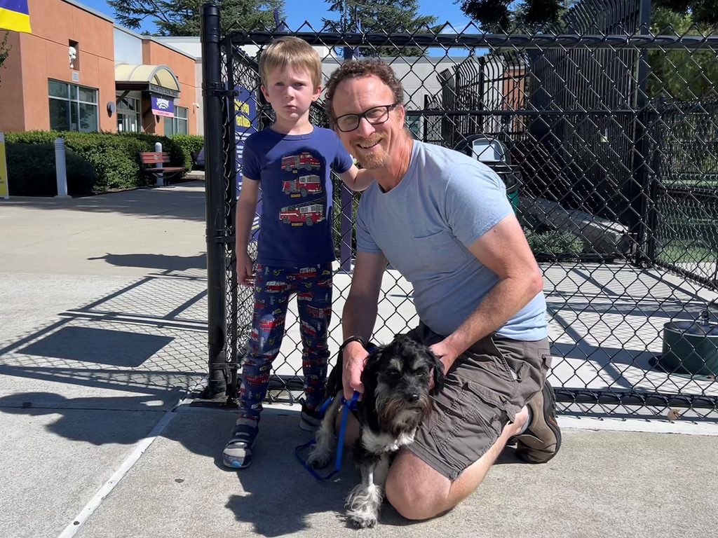 A man kneels beside a small black-and-white dog on a blue leash, with a child standing nearby in front of a chain-link fence outside a building.