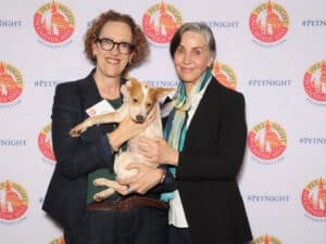 Two women in dark blazers hold a small tan-and-white dog in front of a Pet Night on Capitol Hill backdrop.
