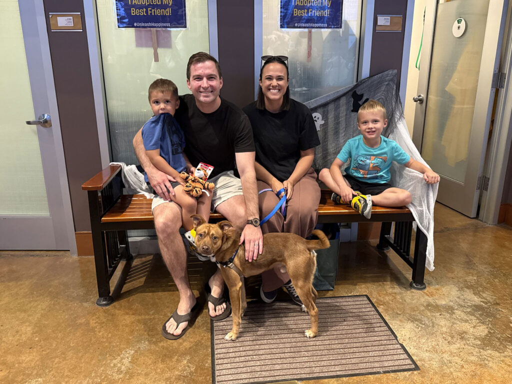 A family with two children sits on a bench with a rescued dog in a shelter lobby, with adoption signs visible on the glass door behind them.