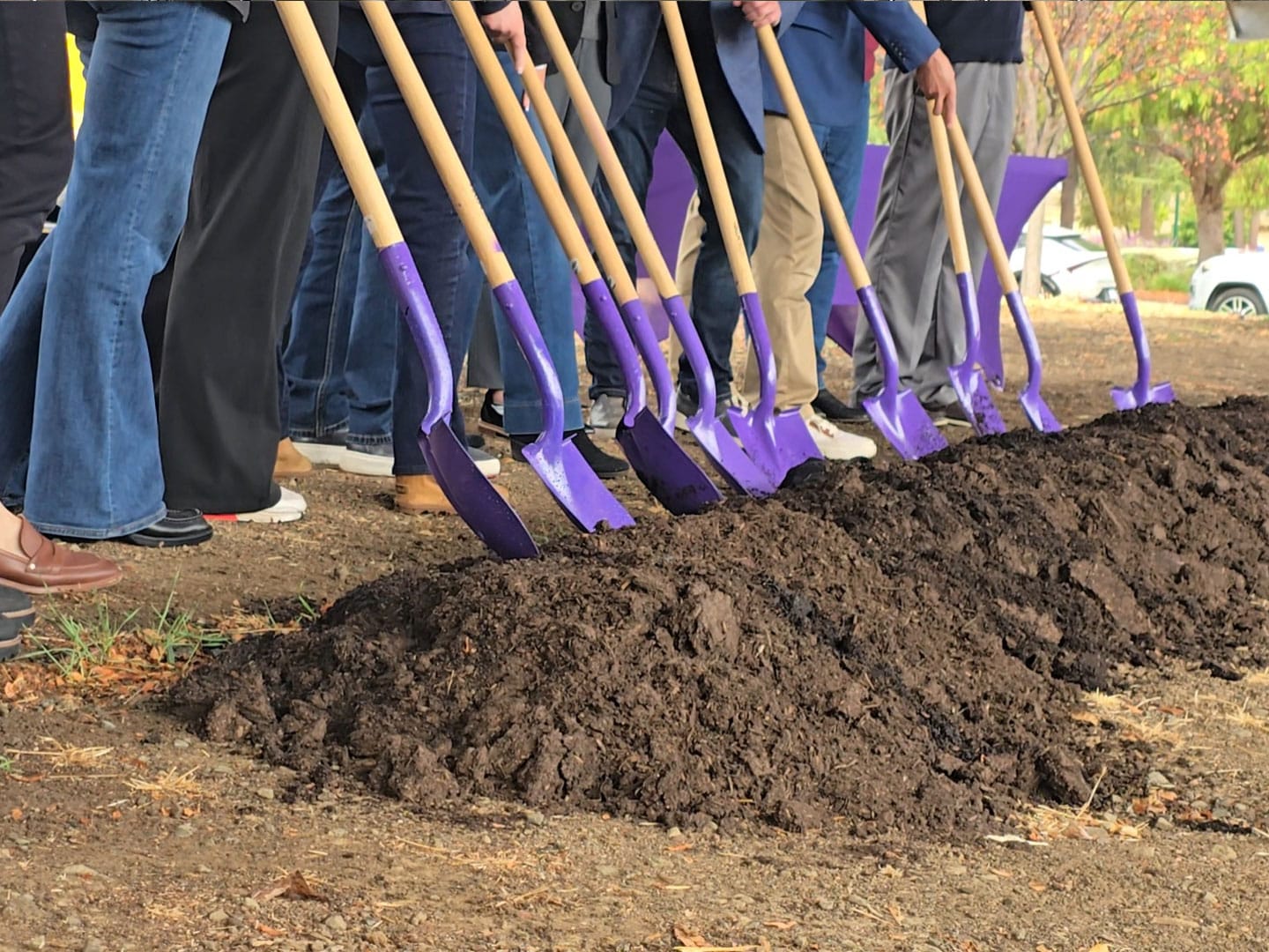 Several people stand in a row holding purple ceremonial shovels beside a long mound of dirt at a groundbreaking event.