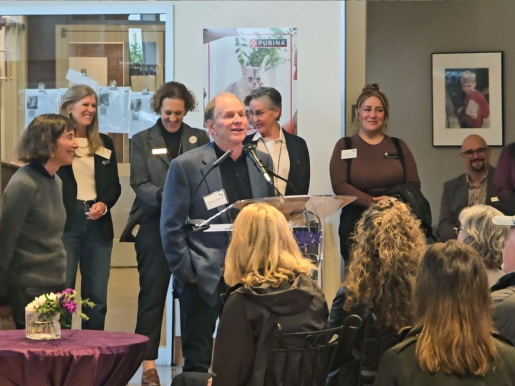 A man speaks at a clear podium during a Liberty Center groundbreaking event, with several people standing behind him and seated attendees in the foreground.