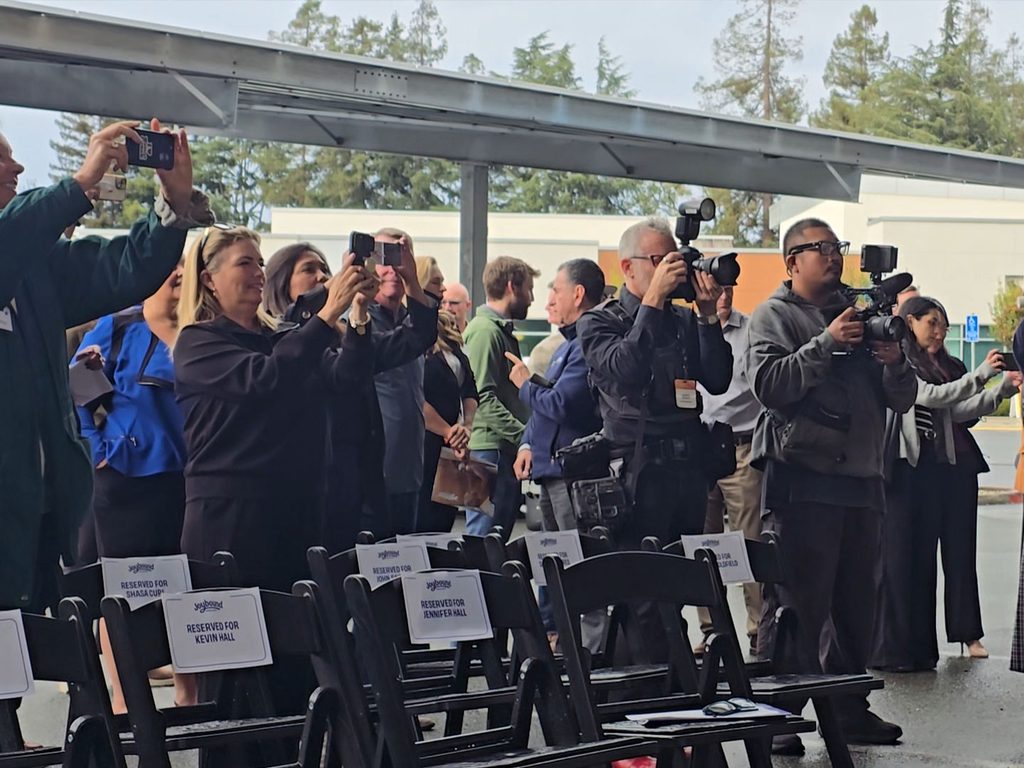 Photographers and attendees stand behind rows of empty black chairs, several holding cameras and smartphones under a covered outdoor area with a roof and trees in the background.