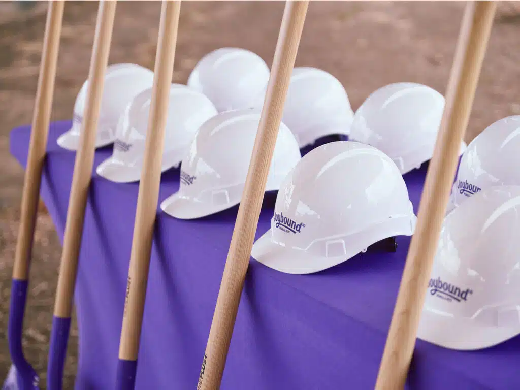 White hard hats lined up on a purple-covered table with wooden-handled shovels in front, prepared for a groundbreaking ceremony.
