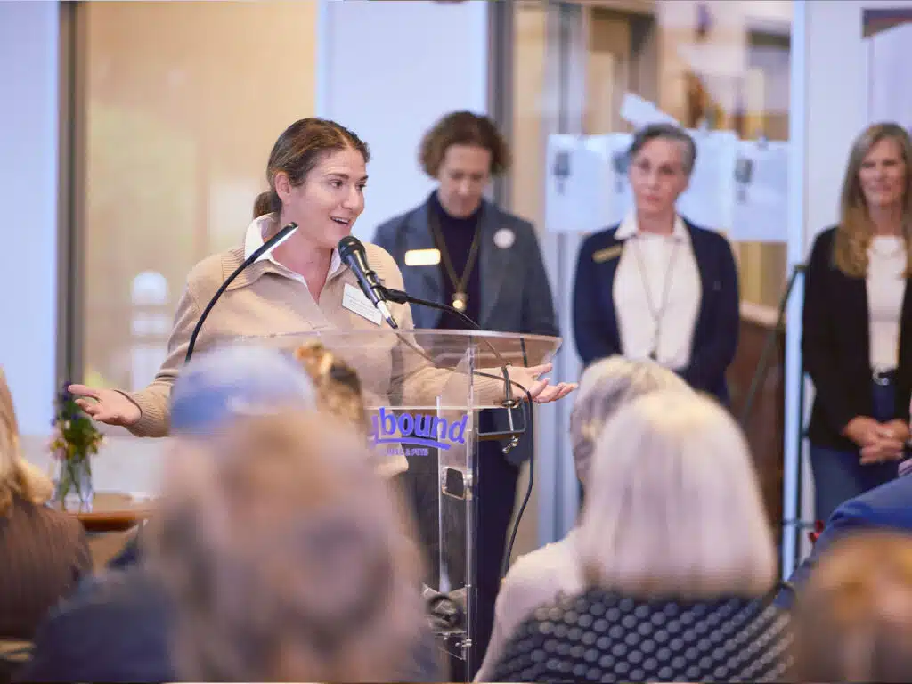 A woman speaks at a podium during a groundbreaking ceremony, with attendees seated in the foreground and three women standing behind her in a lobby.