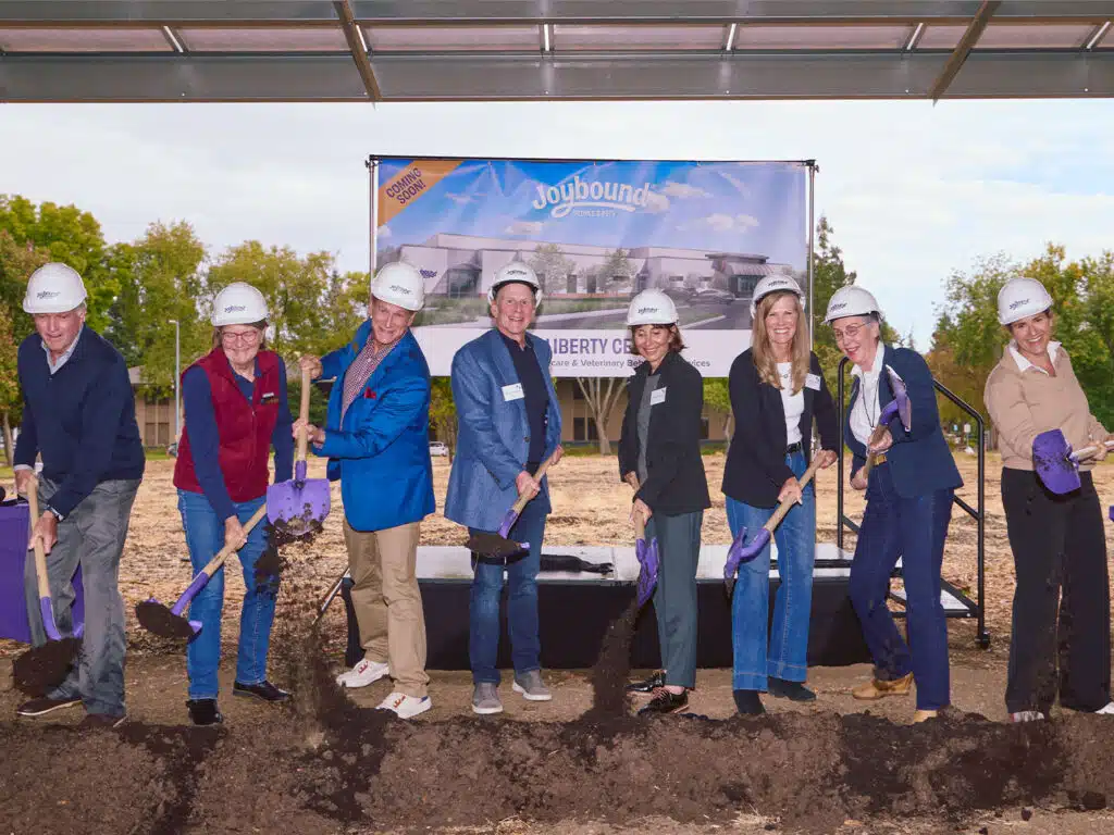 Eight people wearing hard hats stand with purple shovels at a groundbreaking ceremony, with a Liberty Center banner and a dirt area under a shelter behind them.