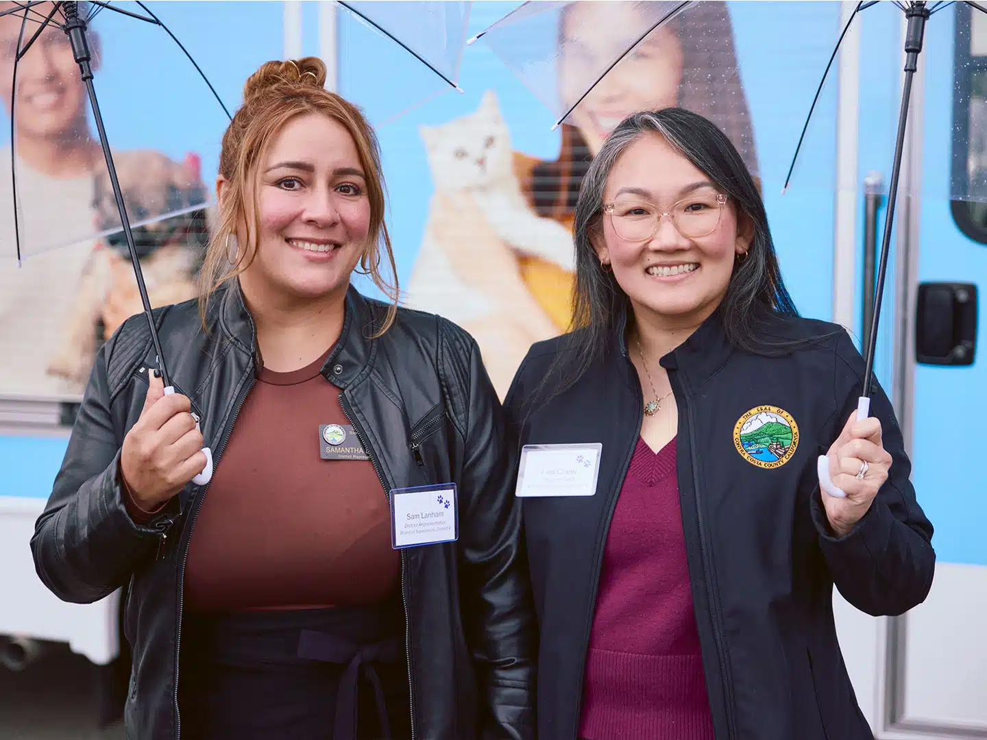 Two women wearing jackets and name badges stand side by side holding clear umbrellas in front of a blue vehicle with a cat graphic.