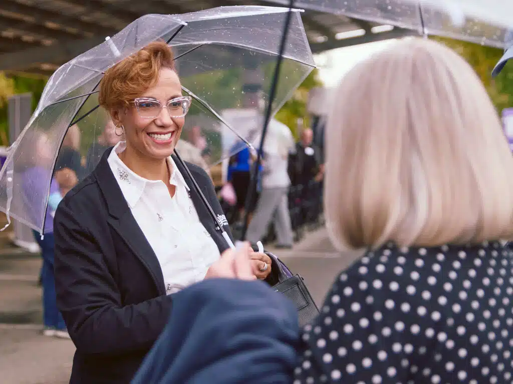A smiling woman in a black blazer and clear umbrella speaks with another woman in a polka-dot top at an outdoor event, with people and a covered walkway in the background.
