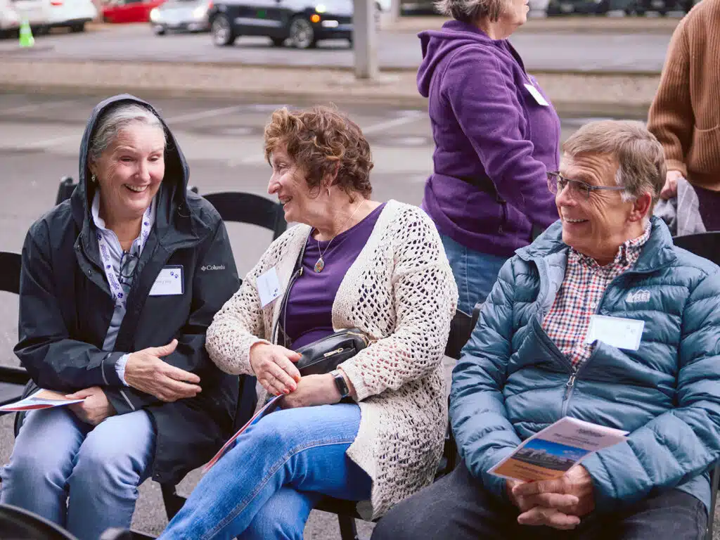 Three older women seated outdoors at a groundbreaking ceremony, including one in a black hooded jacket, one in a white crocheted cardigan, and one in a teal puffer jacket.