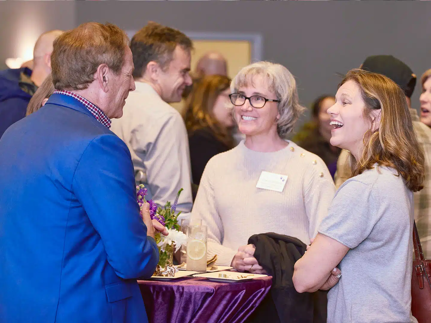 Three adults stand and talk at a reception table with a purple tablecloth, with one man in a blue blazer facing two women in a gray sweater and a light top.