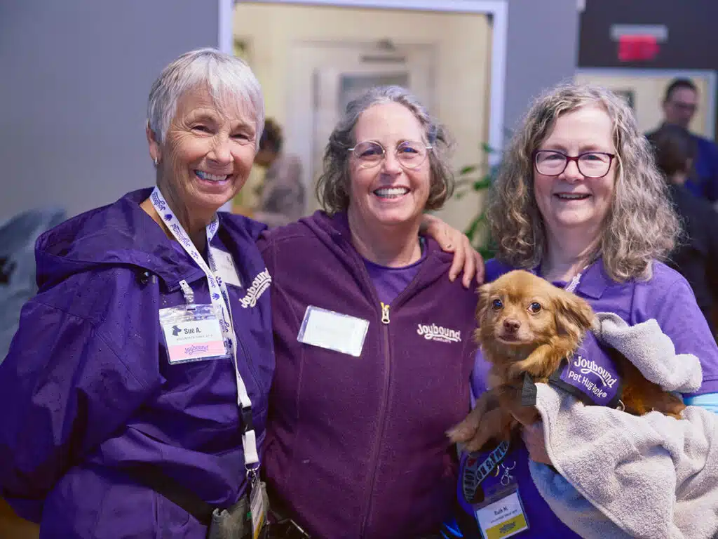 Three women wearing purple tops stand together indoors, with one holding a small brown dog in a purple vest and a blanket.