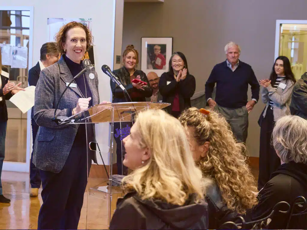 A woman speaks at a clear podium during a groundbreaking ceremony, with applauding attendees standing behind her in a room with framed wall art.