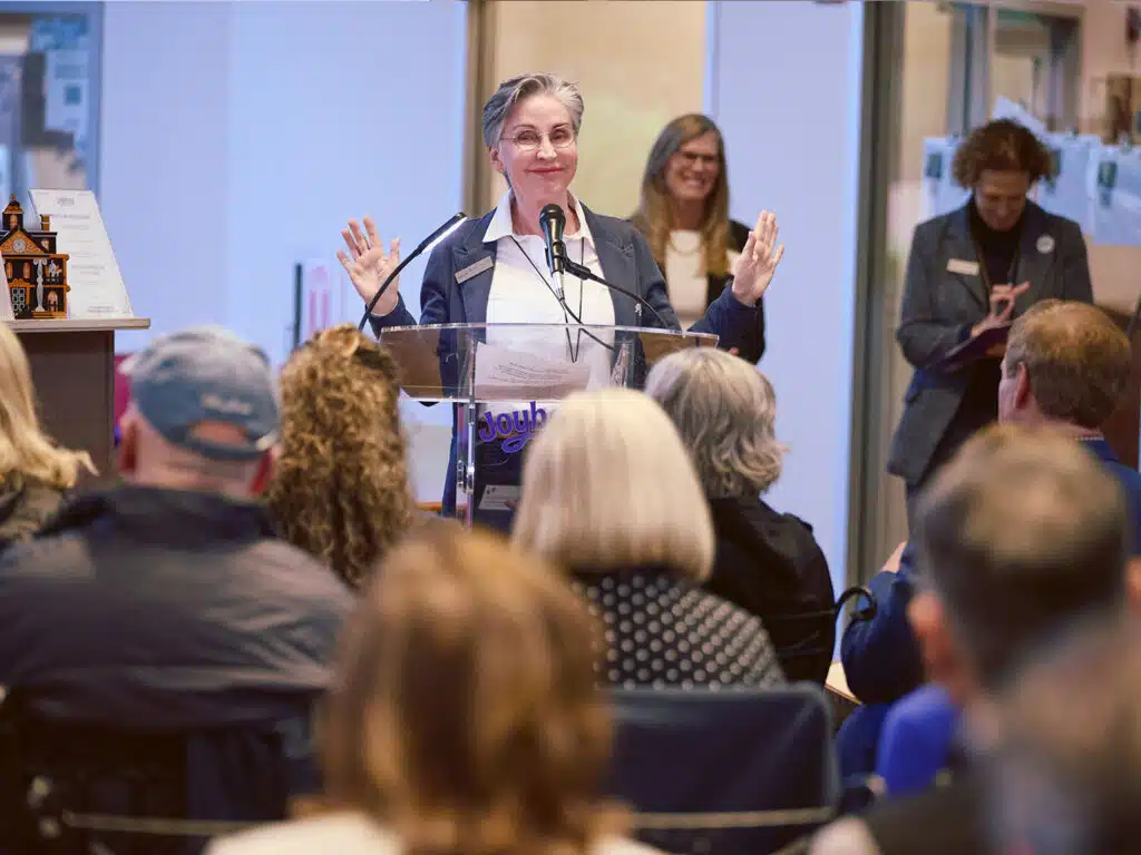 A woman speaks at a clear podium with a microphone, raising both hands, while attendees sit facing her in a bright indoor venue.