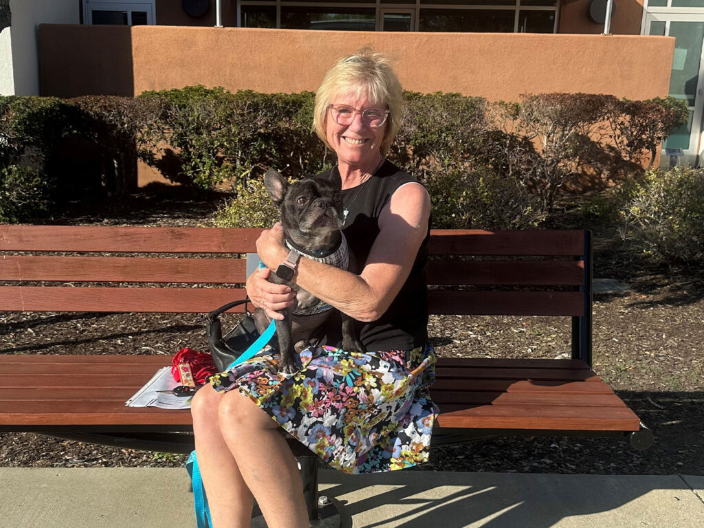 A smiling woman in glasses sits on a wooden bench holding a small black dog on a blue leash, with hedges and a building behind her.
