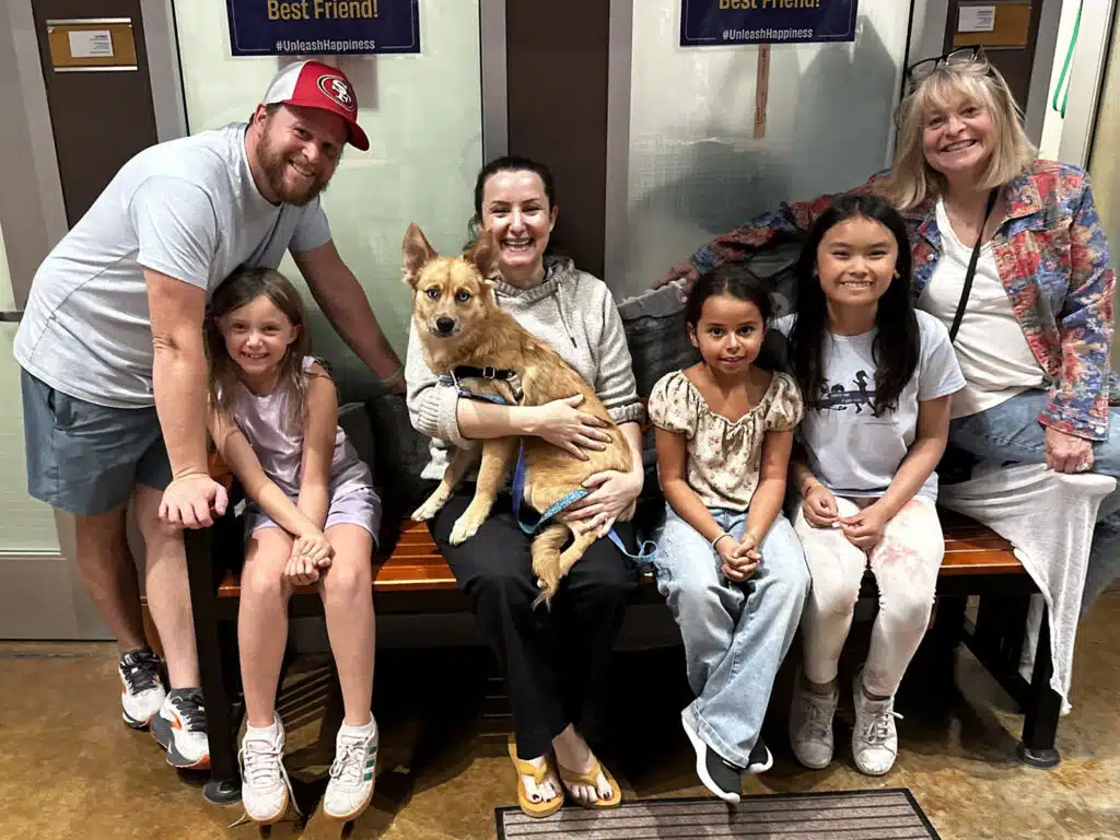 A family of six sits on a bench with a tan dog in a pet adoption setting, in front of a sign that reads Best Friend!