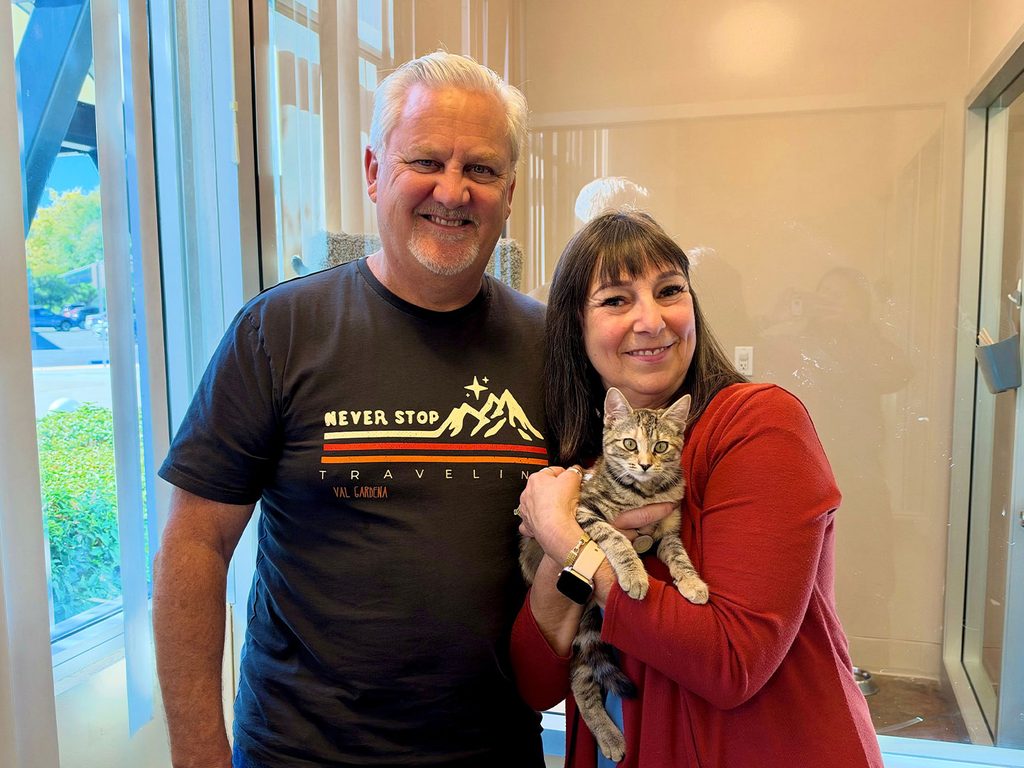 A smiling couple stands indoors beside a tabby kitten held in the woman’s arms, with a doorway and windowed hallway in the background.