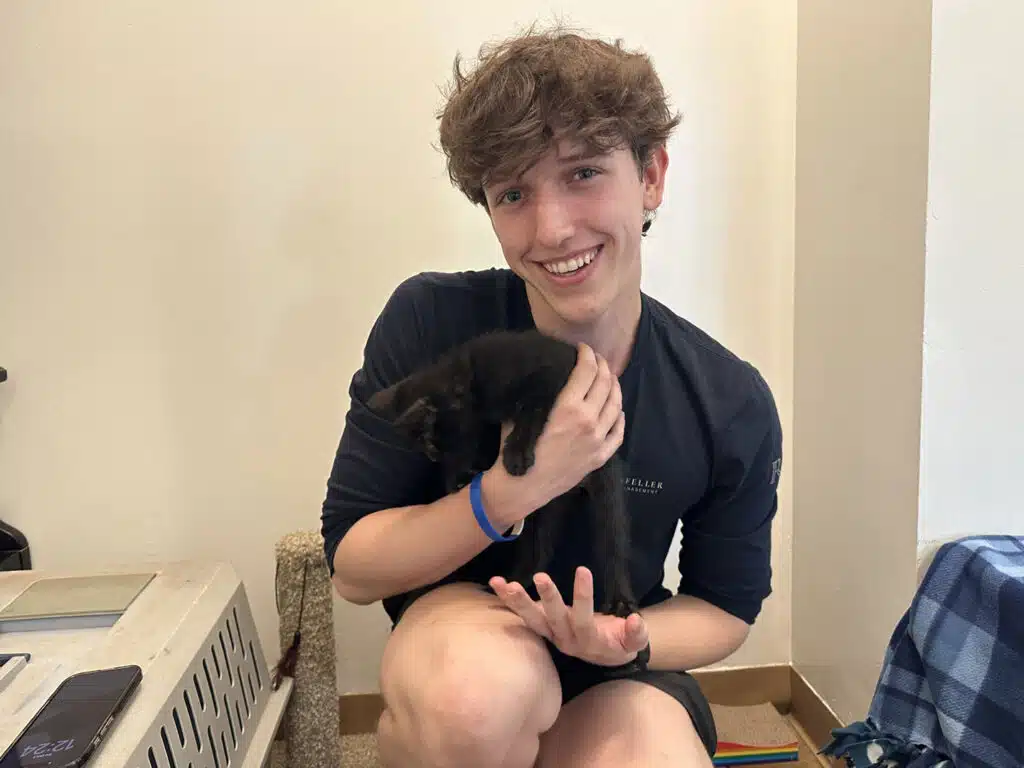 A young man in a dark shirt holds a small black kitten indoors beside a pet carrier and a blue blanket.
