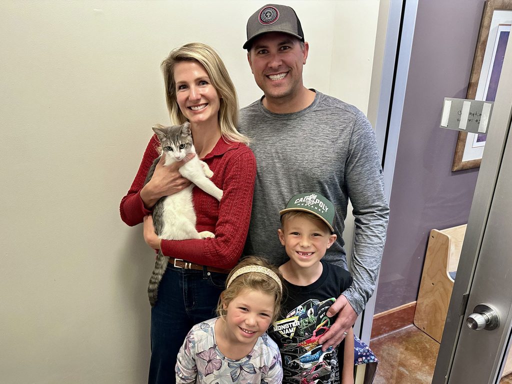 A family of four stands indoors holding a gray-and-white cat, with a hallway and door visible behind them.