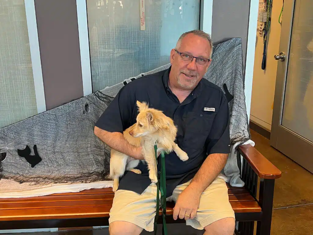 A man in glasses and a navy shirt sits on a wooden bench holding a small tan-and-white dog on a green leash, with a gray blanket draped across the bench and a door in the background.