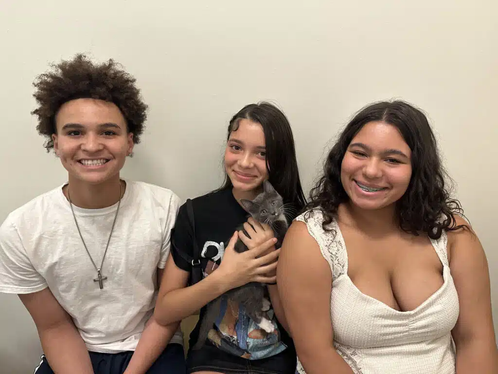 Three teenagers sit against a plain wall, with the center teen holding a gray kitten on her lap.