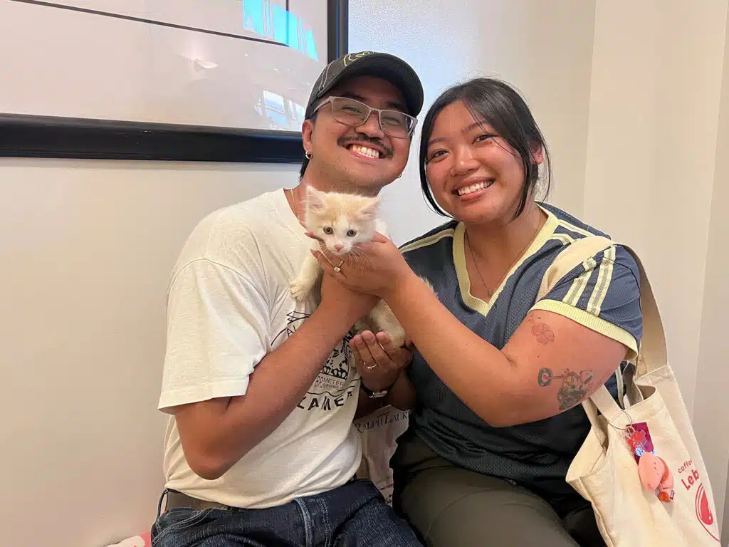 A smiling couple holds a small cream-colored kitten indoors, with a framed picture on the wall behind them.