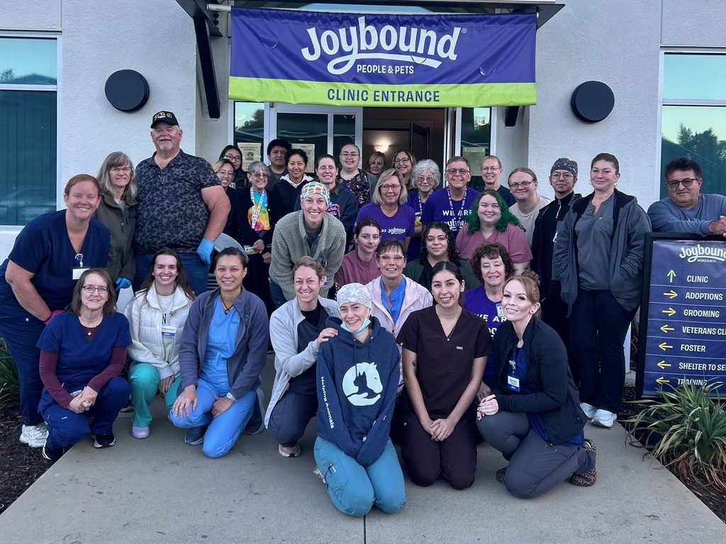 A group of people pose outside the Joybound People & Pets clinic entrance, with a blue-and-green banner overhead and a sign listing clinic and adoption directions nearby.