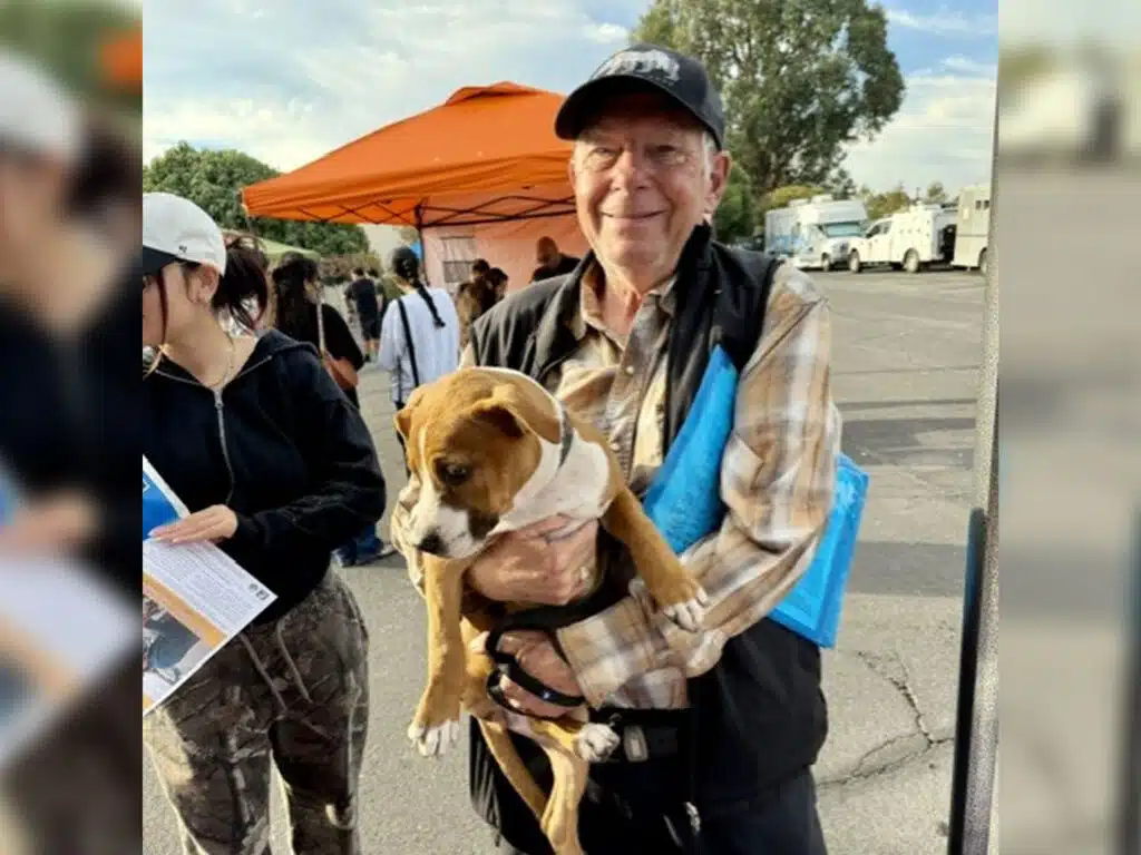 An older man in a black cap holds a brown-and-white puppy at an outdoor food pantry, with an orange canopy and other people in the background.