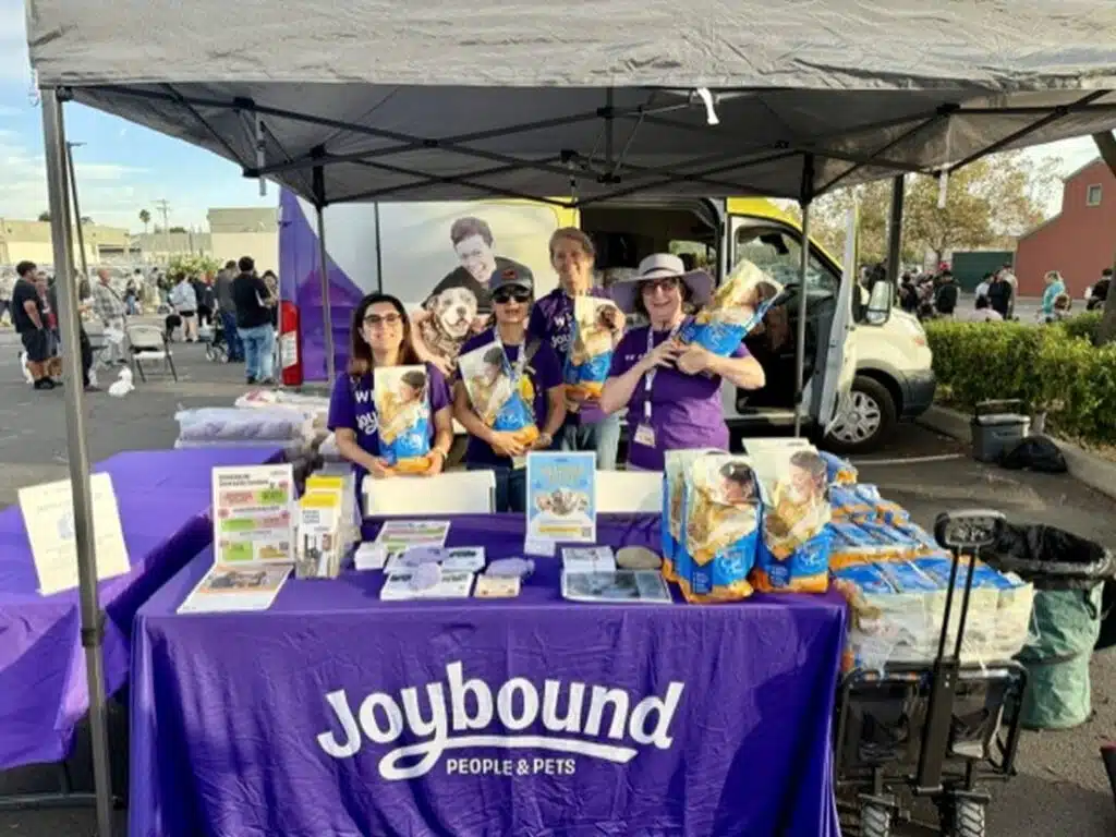 Four volunteers stand behind a Joybound People & Pets table under a black canopy, with flyers, pet food bags, and stackable display boards at an outdoor event.