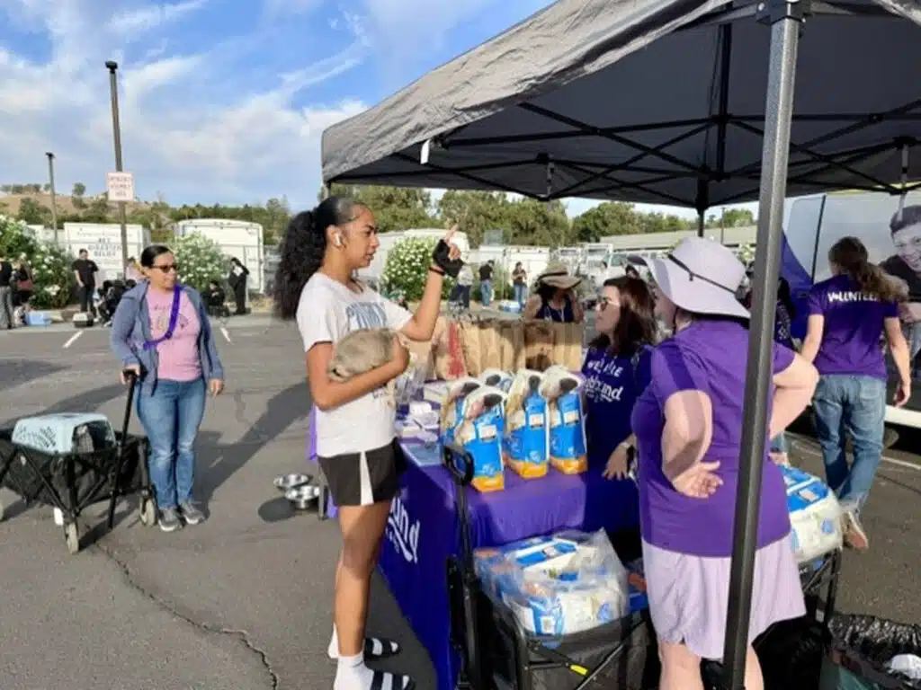 A young woman in a white T-shirt stands by a canopy table stocked with food bags and bottled water, with volunteers in purple shirts and people gathered in the parking lot behind her.