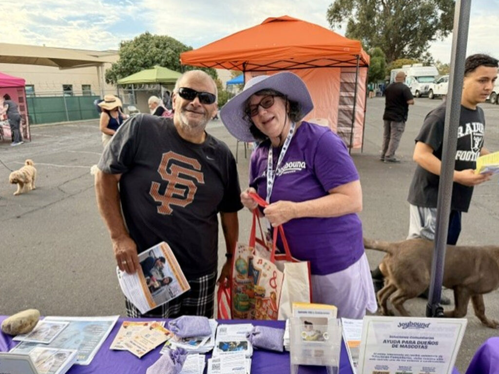 A smiling man in a black San Francisco Giants shirt stands beside a woman in a purple hat and shirt at a FoodShare Pantry table, with brochures, tote bags, and dogs visible in the background.