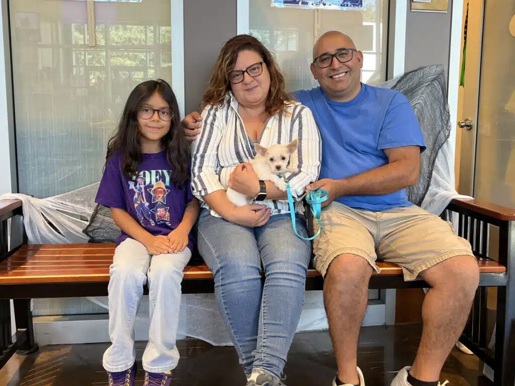 A family of three sits on a bench holding a small white dog with a blue leash, in front of large windows.