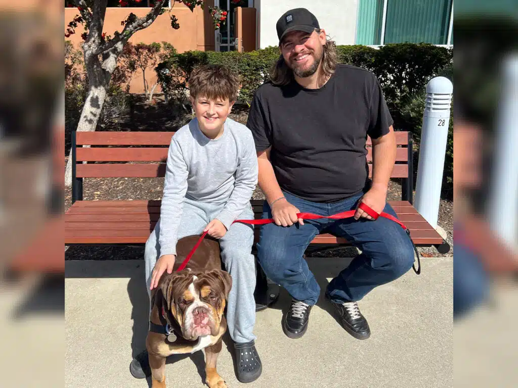 A boy and a man sit on a bench holding a bulldog on a red leash in an outdoor area with a tree, shrubs, and a building behind them.