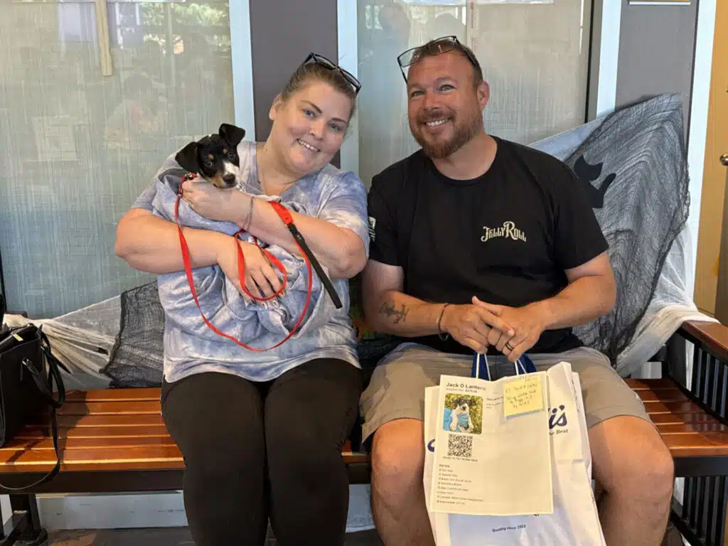 A woman holding a small black-and-white dog sits beside a man on a bench in an indoor waiting area, with adoption paperwork and a tote bag on his lap.