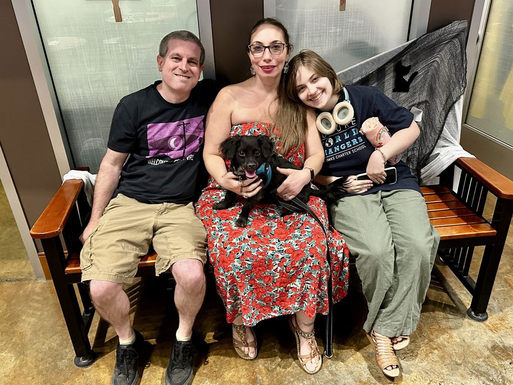 A man, woman, and child sit on a wooden bench holding a small black puppy, with a frosted glass wall behind them.