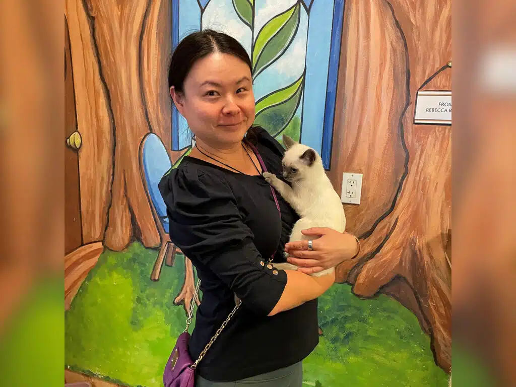 A woman holding a Siamese kitten stands in front of a painted forest mural, with a wall sign and electrical outlet visible beside her.