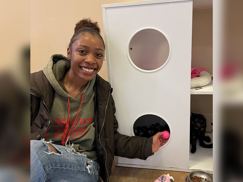 A smiling woman kneels beside a white cat shelter unit with circular openings, holding a pink toy near a black cat inside a cubby.
