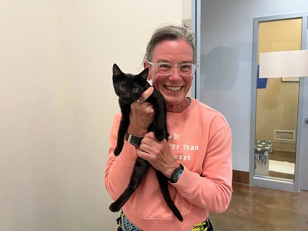 A smiling older woman in a coral sweatshirt holds a black cat, with an indoor hallway and glass door behind her.