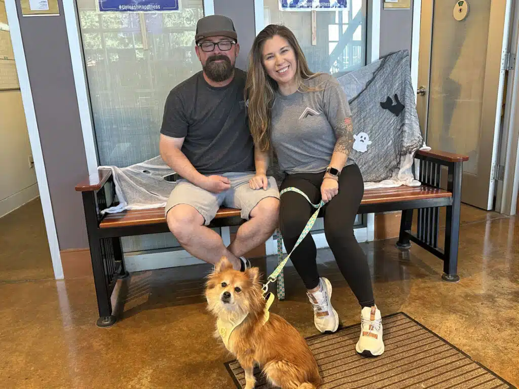 A smiling couple sits on a bench with a small brown dog on a leash in the foreground, inside a pet adoption area with a doorway and floor mat.