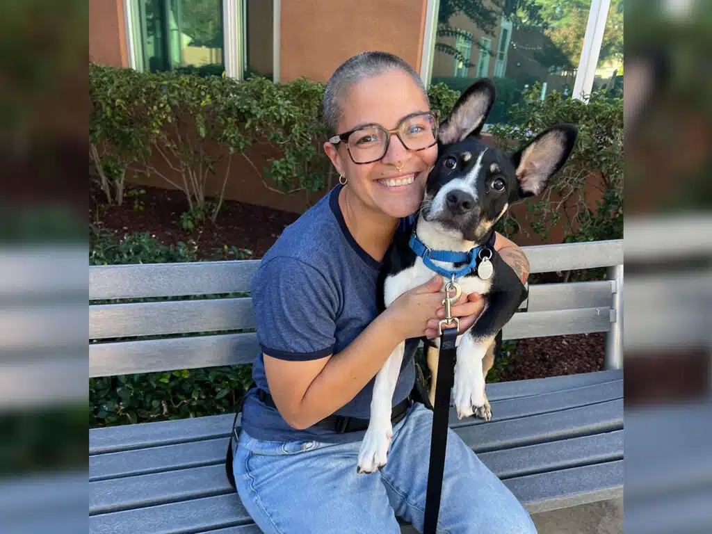 A smiling woman with glasses sits on a bench holding a black-and-white dog with a blue collar, with shrubs and a building in the background.