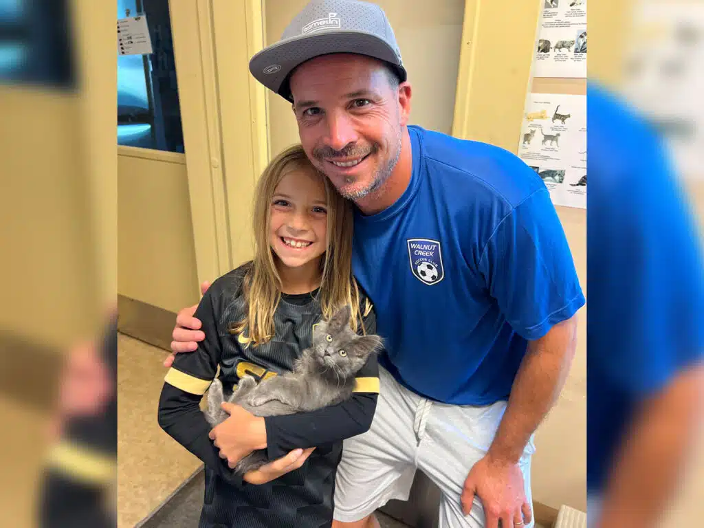 A man in a gray cap and blue shirt poses with a smiling girl holding a gray kitten, standing indoors near a doorway and pet adoption posters.