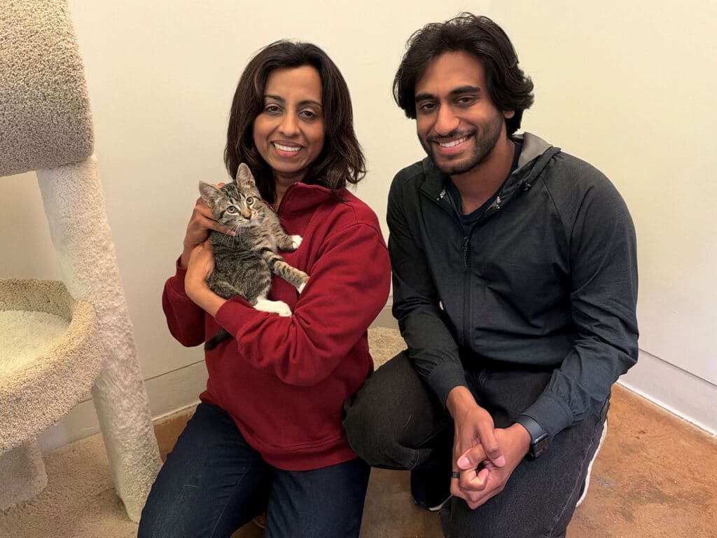 A woman in a red jacket holds a tabby cat while a man kneels beside her, with a beige cat tree and plain wall in the background.