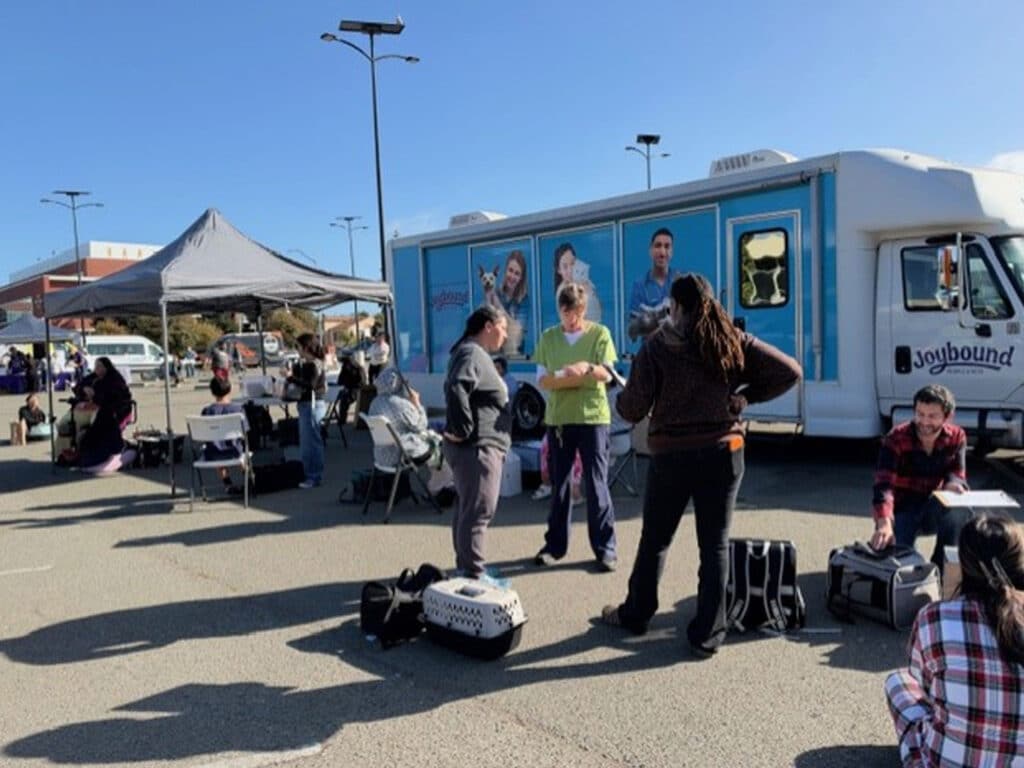 People gather beside a mobile clinic truck and tented food pantry in a parking lot under a clear blue sky.