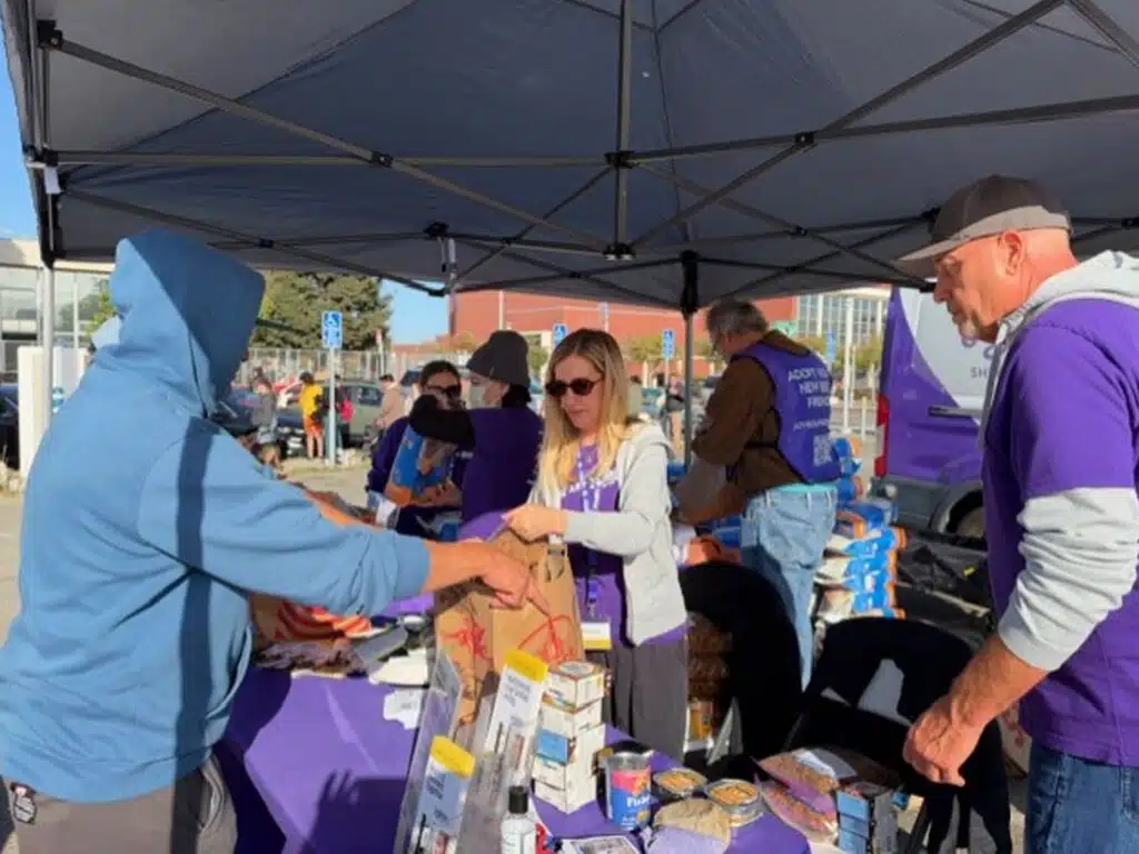 People under a canopy exchange groceries and supplies at an outdoor Vaccine Clinic and FoodShare Pantry table.